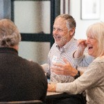 Sitting at a table, a man gestures while a woman laughs